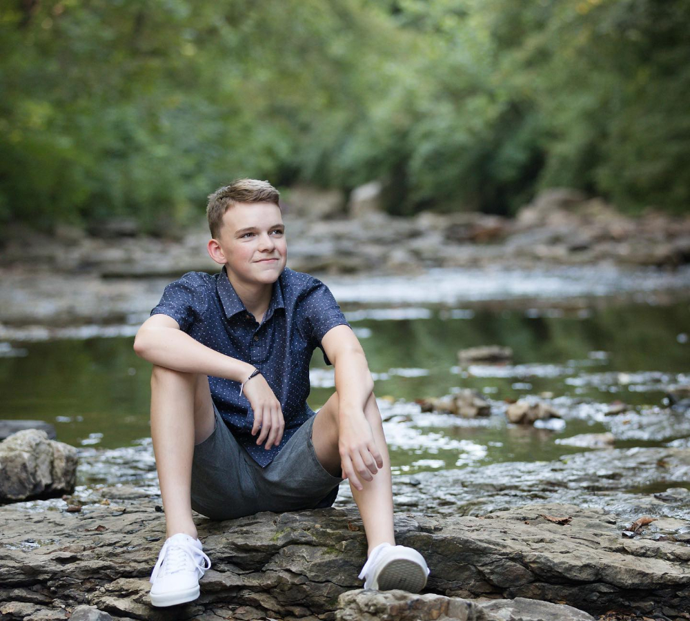 Young person in a blue shirt sits relaxed on rocks by a stream, surrounded by lush greenery, exuding a calm and thoughtful mood.