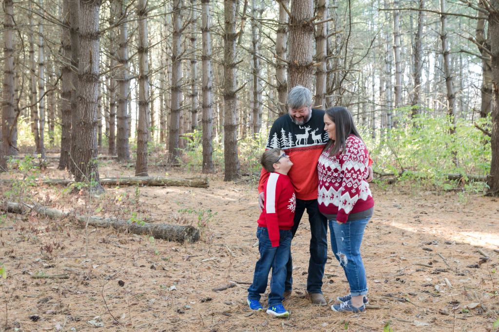 Family in festive sweaters smiles in a pine forest. Boy in red looks up at adults, surrounded by tall trees and scattered logs. Cozy mood.