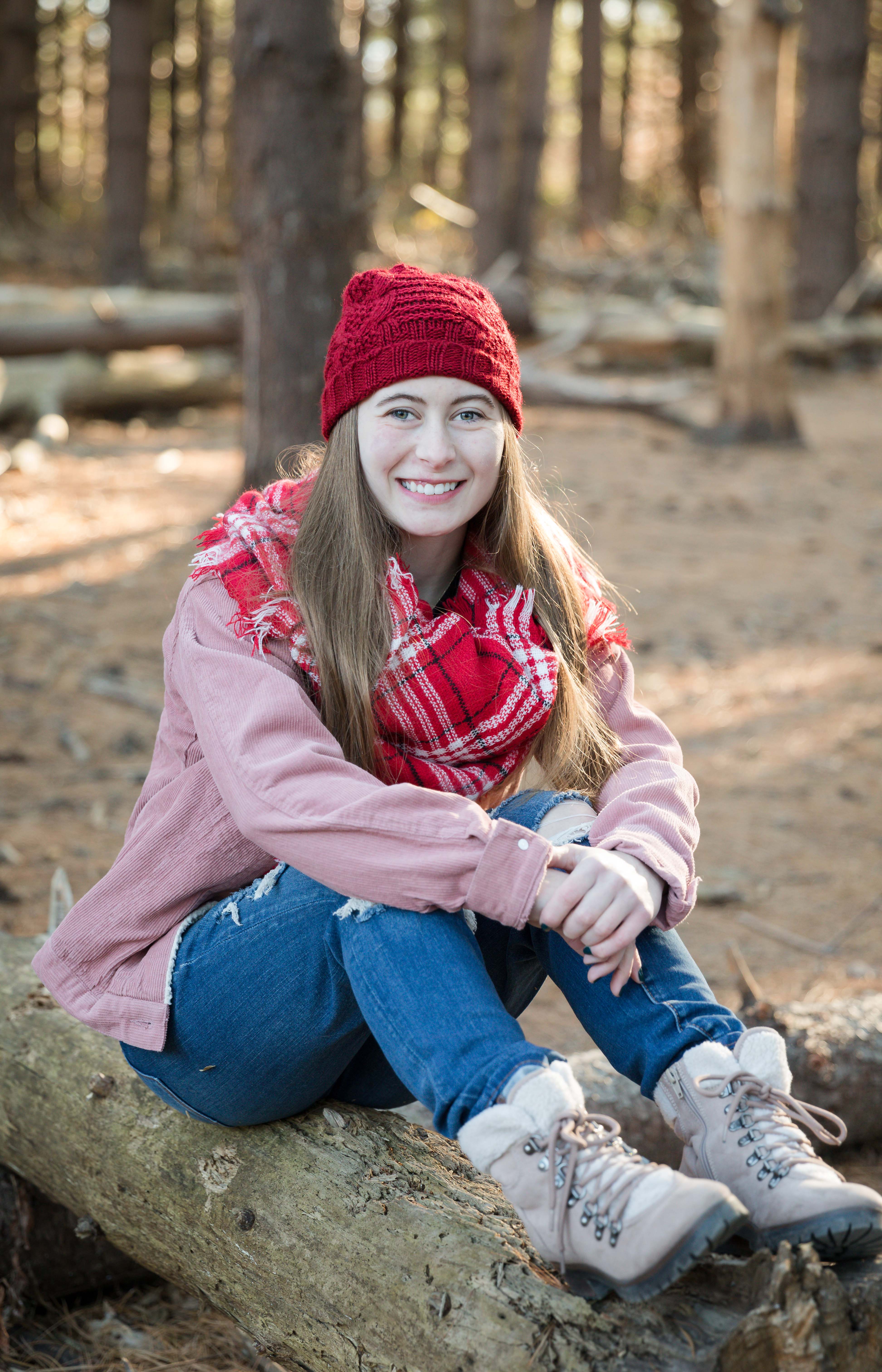 Smiling woman in red hat and plaid scarf sits on a log in a sunlit forest, wearing a pink jacket, jeans, and boots. Cozy and cheerful mood.