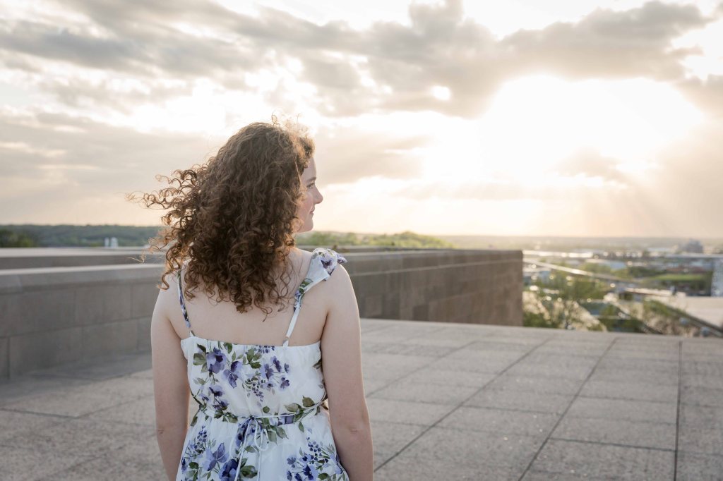 Curly-haired woman in floral dress gazes at sunset on rooftop. Sunlight breaks through clouds. Mood is serene and contemplative.