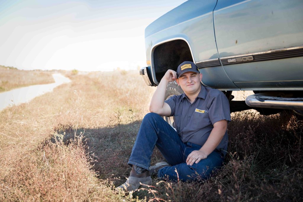 A man in a cap and uniform sits next to a blue GMC 1500 truck in a grassy field, looking relaxed. A dirt road stretches into the distance.
