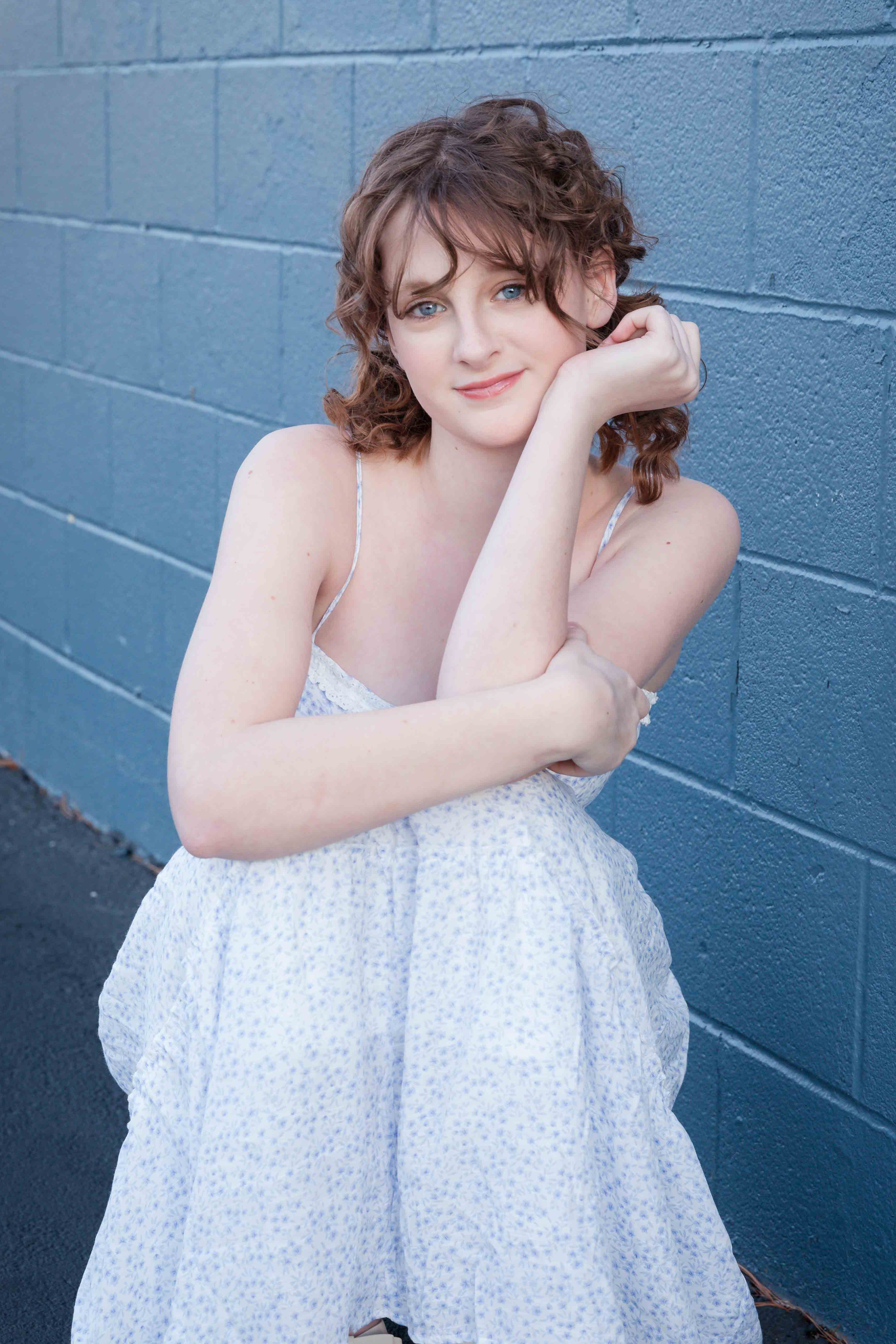 Woman in a white patterned dress sitting against a blue brick wall, smiling softly with one hand on her cheek, creating a serene mood.