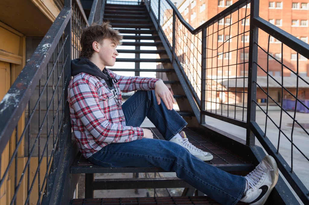 Teen in plaid shirt sits thoughtfully on outdoor metal stairs, urban brick building in background, calm and relaxed mood. Case Studios Photography Senior