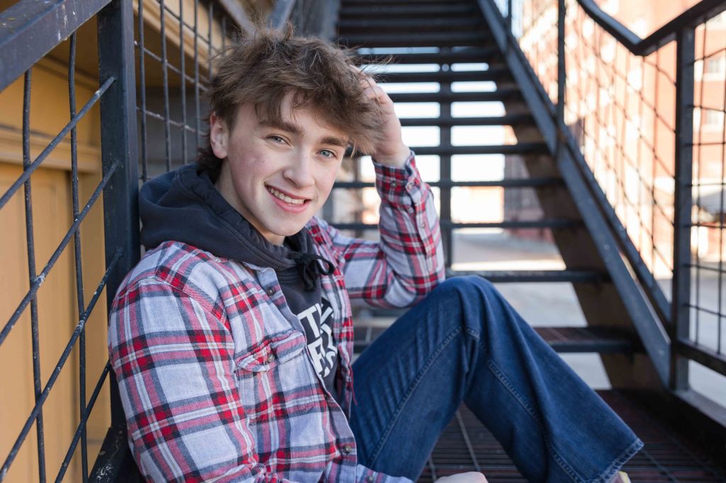Young person in plaid shirt smiles, sitting on outdoor metal stairs, touching hair. Background features railings and a yellow wall. Casual mood.