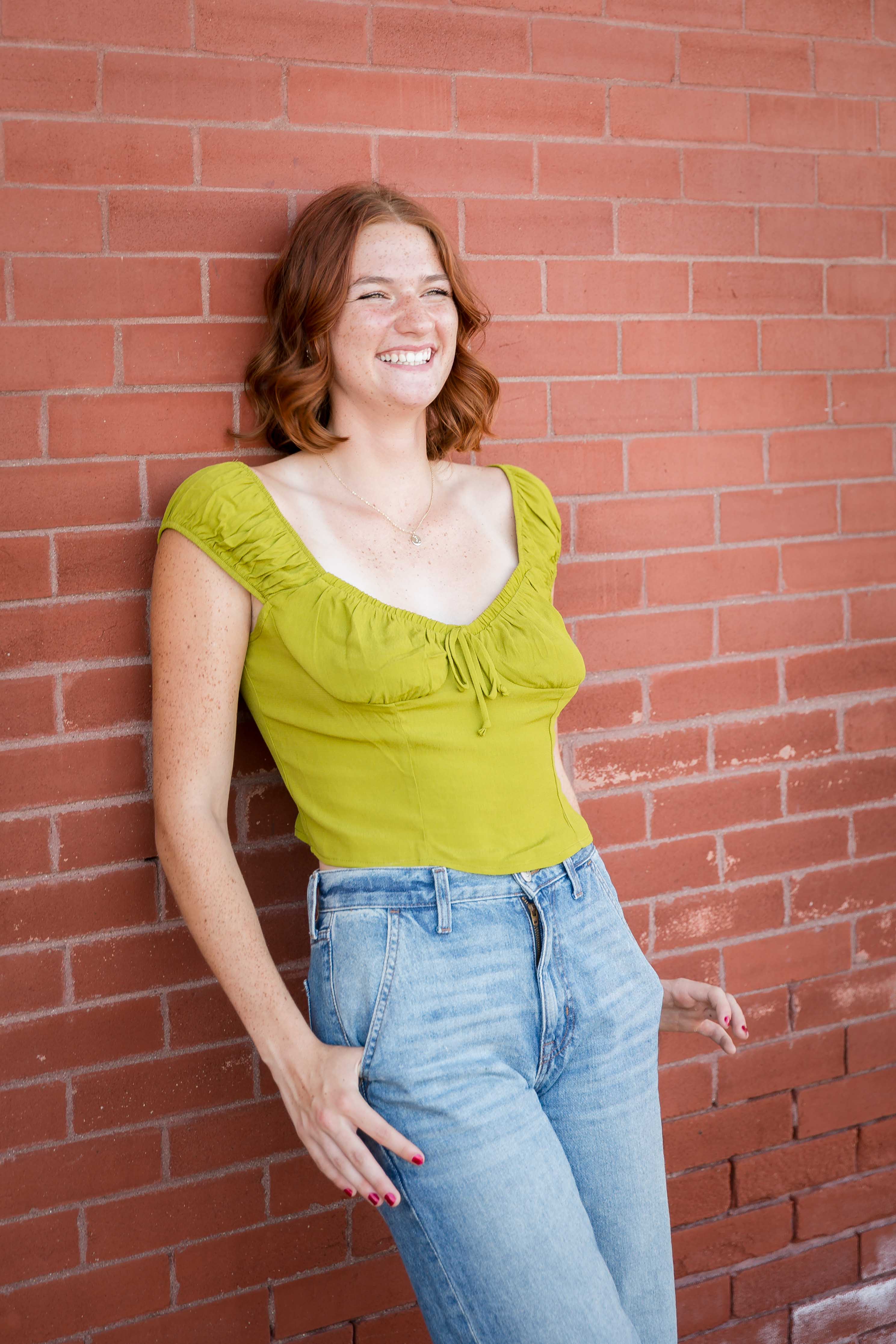 Smiling woman with curly red hair leans against a red brick wall, wearing a green top and jeans. Bright and cheerful mood.