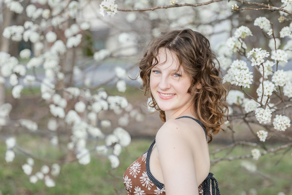Smiling woman with curly hair in floral dress stands near blossoming tree. Bright, sunny setting with white flowers in background.