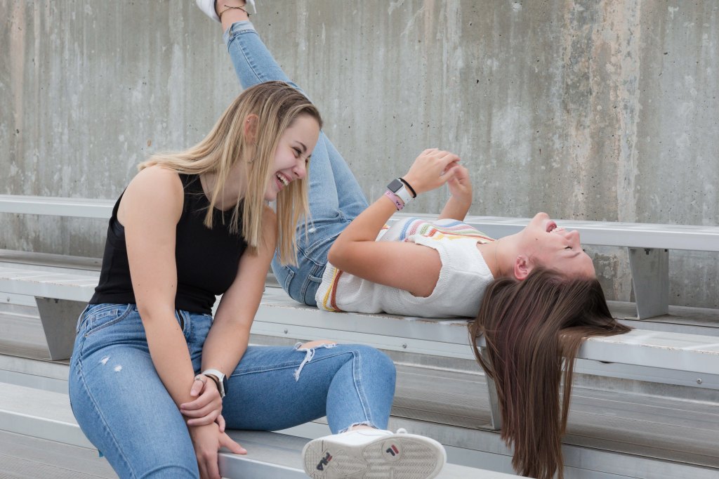 Two women in casual jeans, one sitting and the other lying on bleachers, laughing. Concrete wall in the background adds a casual vibe.