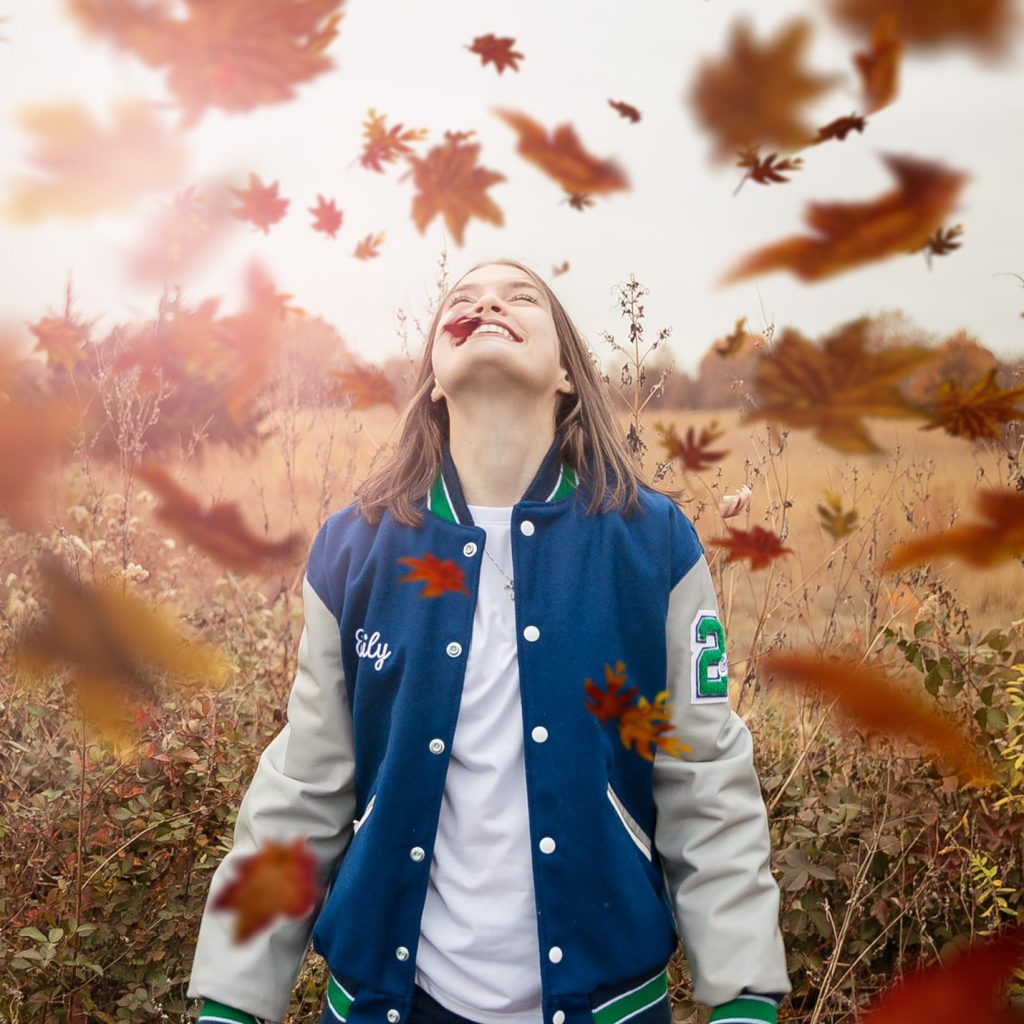 Smiling person in blue jacket stands outdoors, surrounded by falling autumn leaves. Overcast sky and dried plants in background.