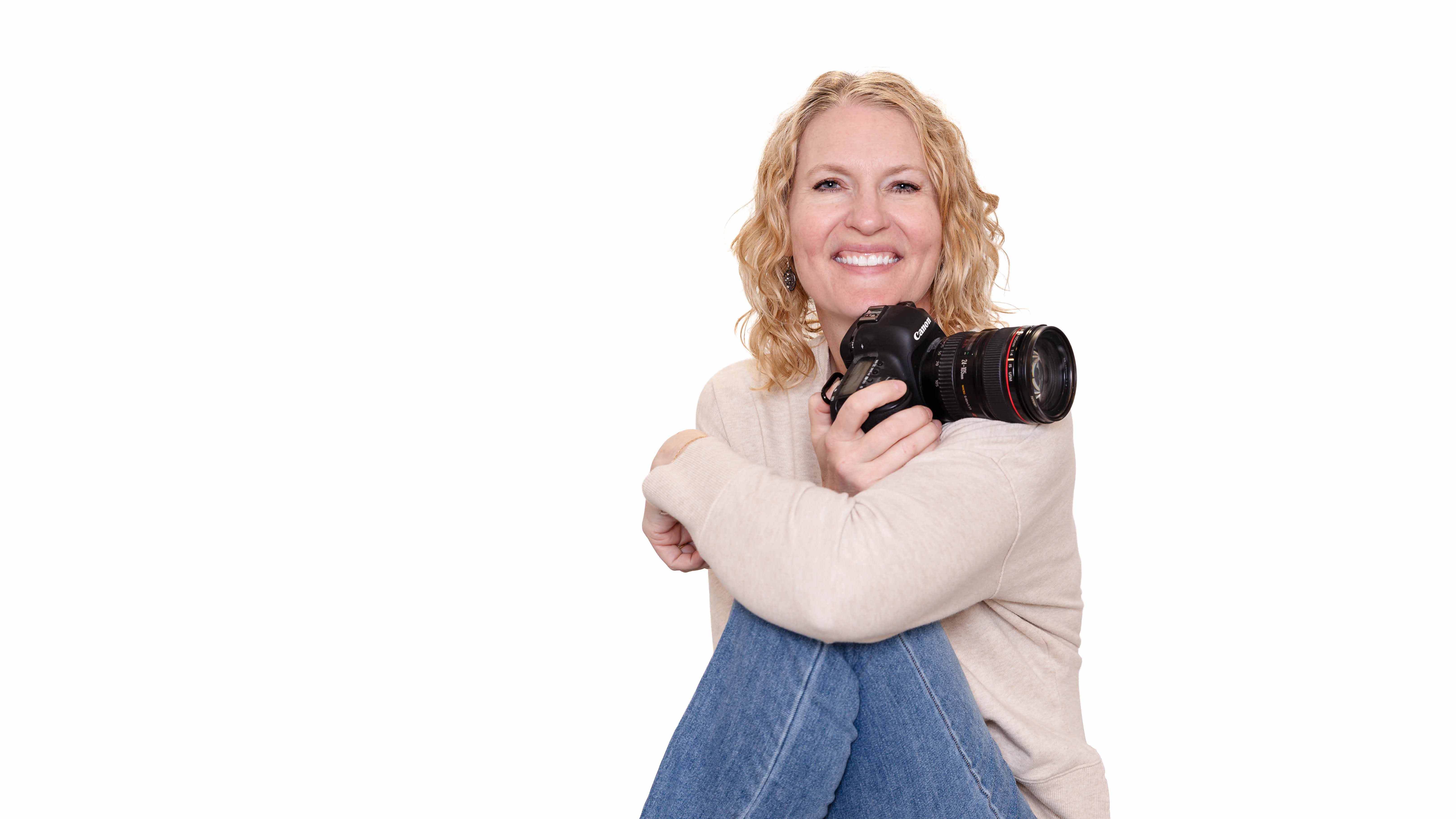 A person with curly hair smiles while holding a camera. They wear a beige sweater with blue text. White background. Mood is joyful.