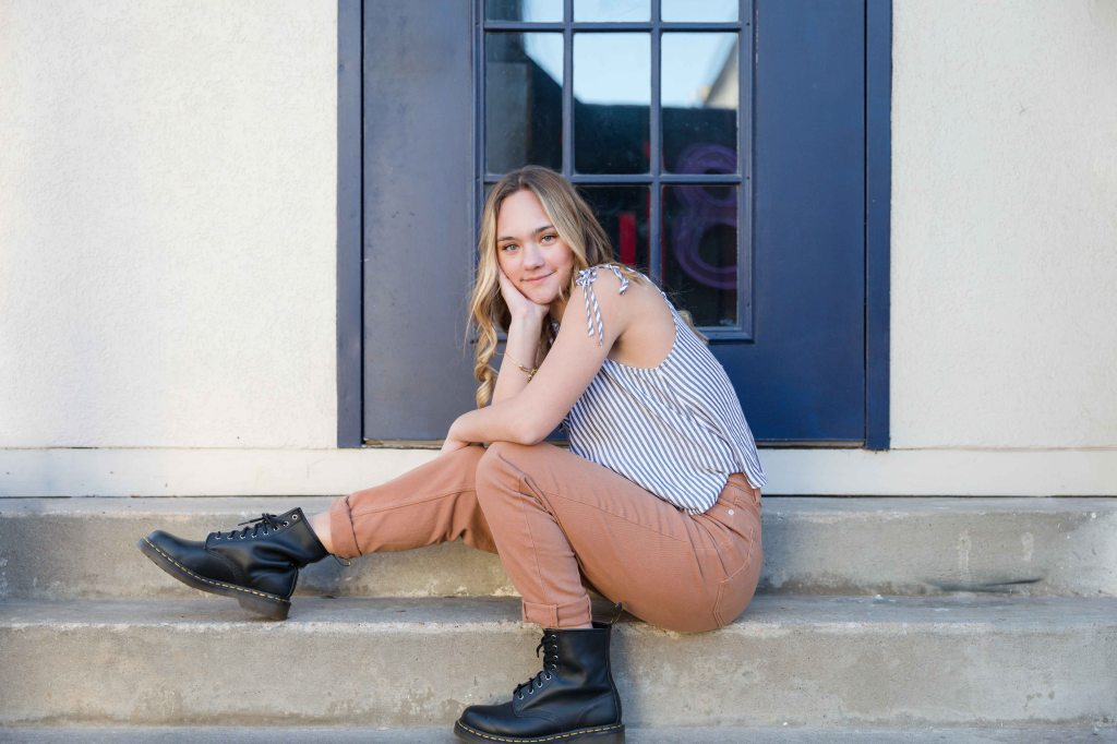 Young woman sits on concrete steps, smiling. She wears a striped top, brown pants, and black boots. Blue door and window in background.