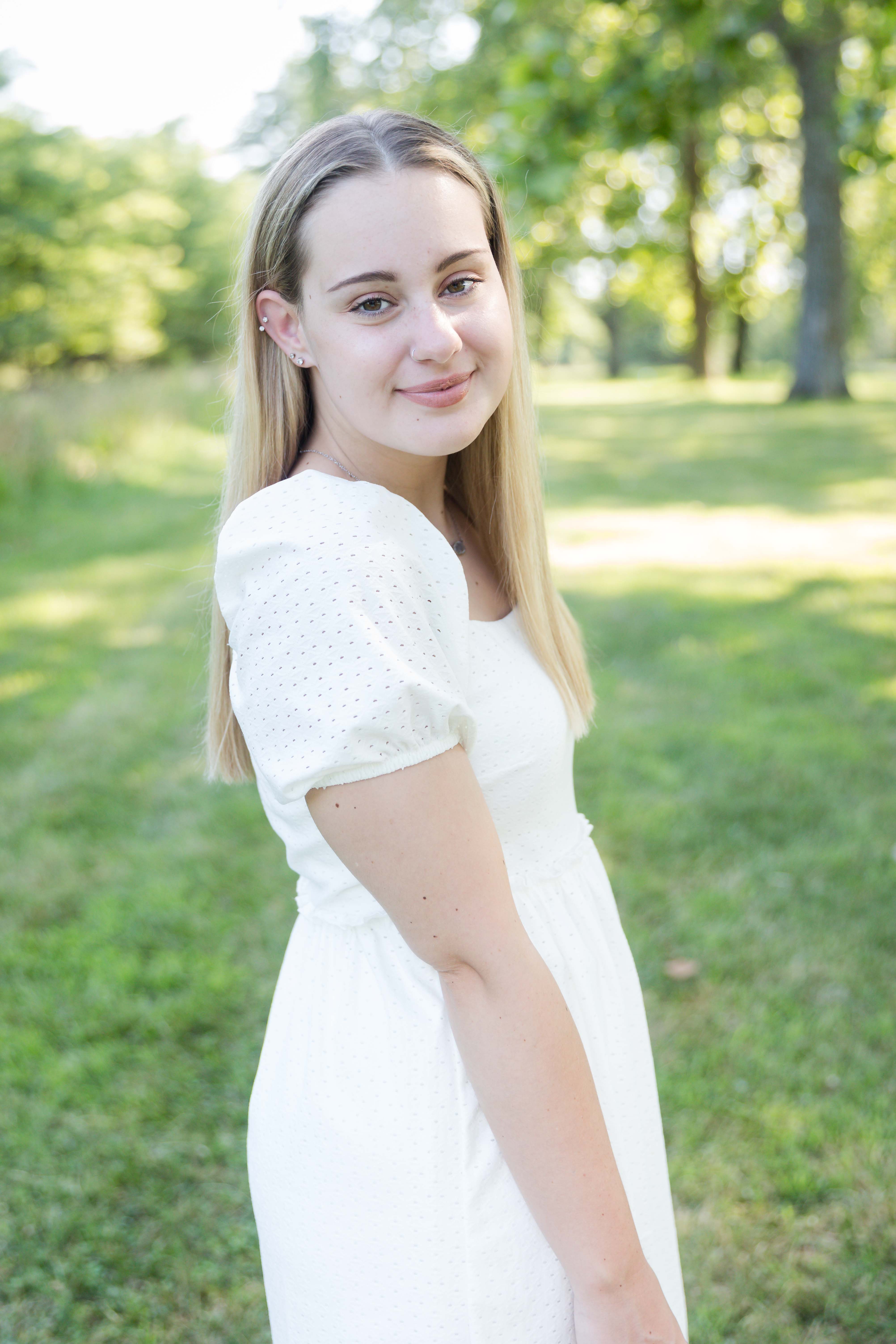 Smiling woman in a white dress stands in a sunlit park with lush green trees, conveying a serene and cheerful mood.