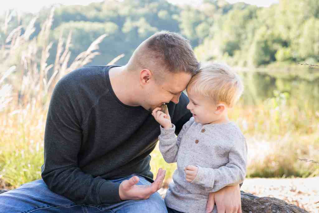 Father and son share a joyful moment, touching foreheads outdoors. Both are smiling, surrounded by greenery and a serene lake in the background.