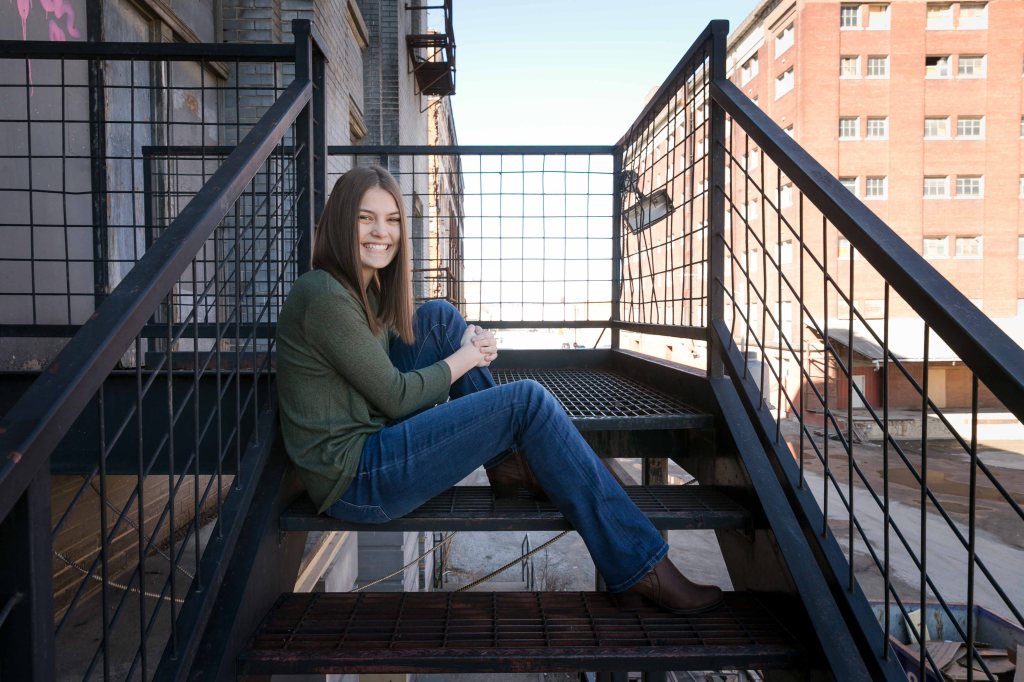 Smiling woman in green sweater and jeans sits on an outdoor metal staircase. Background shows industrial brick buildings and a clear sky.