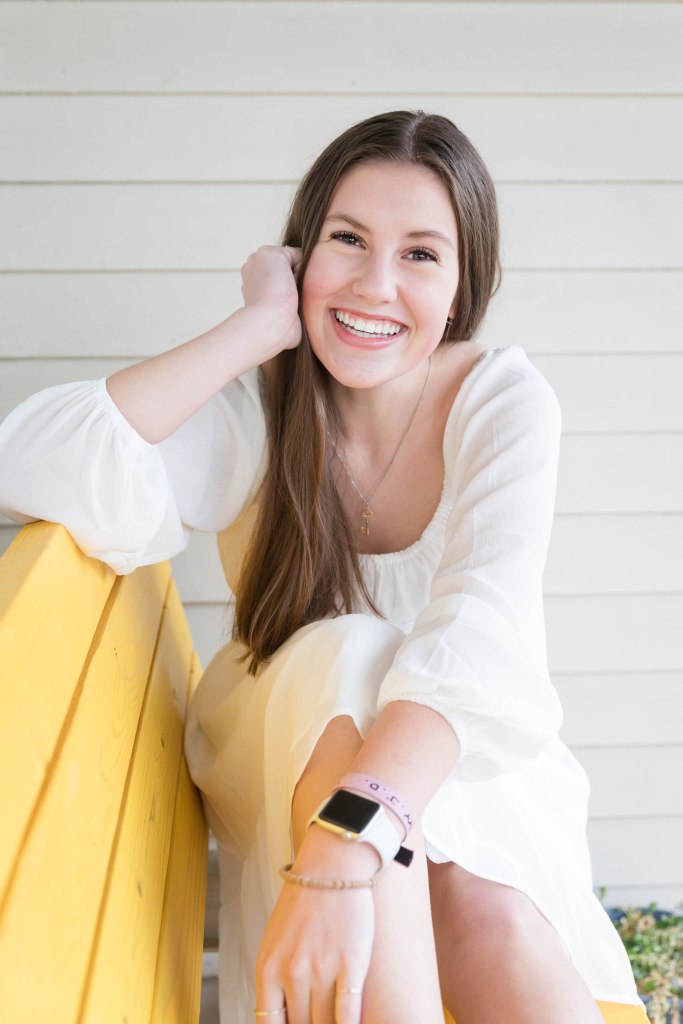 A smiling woman in a white dress sits on a yellow bench, leaning on her hand. She's wearing a smartwatch, with a bright, cheerful expression.