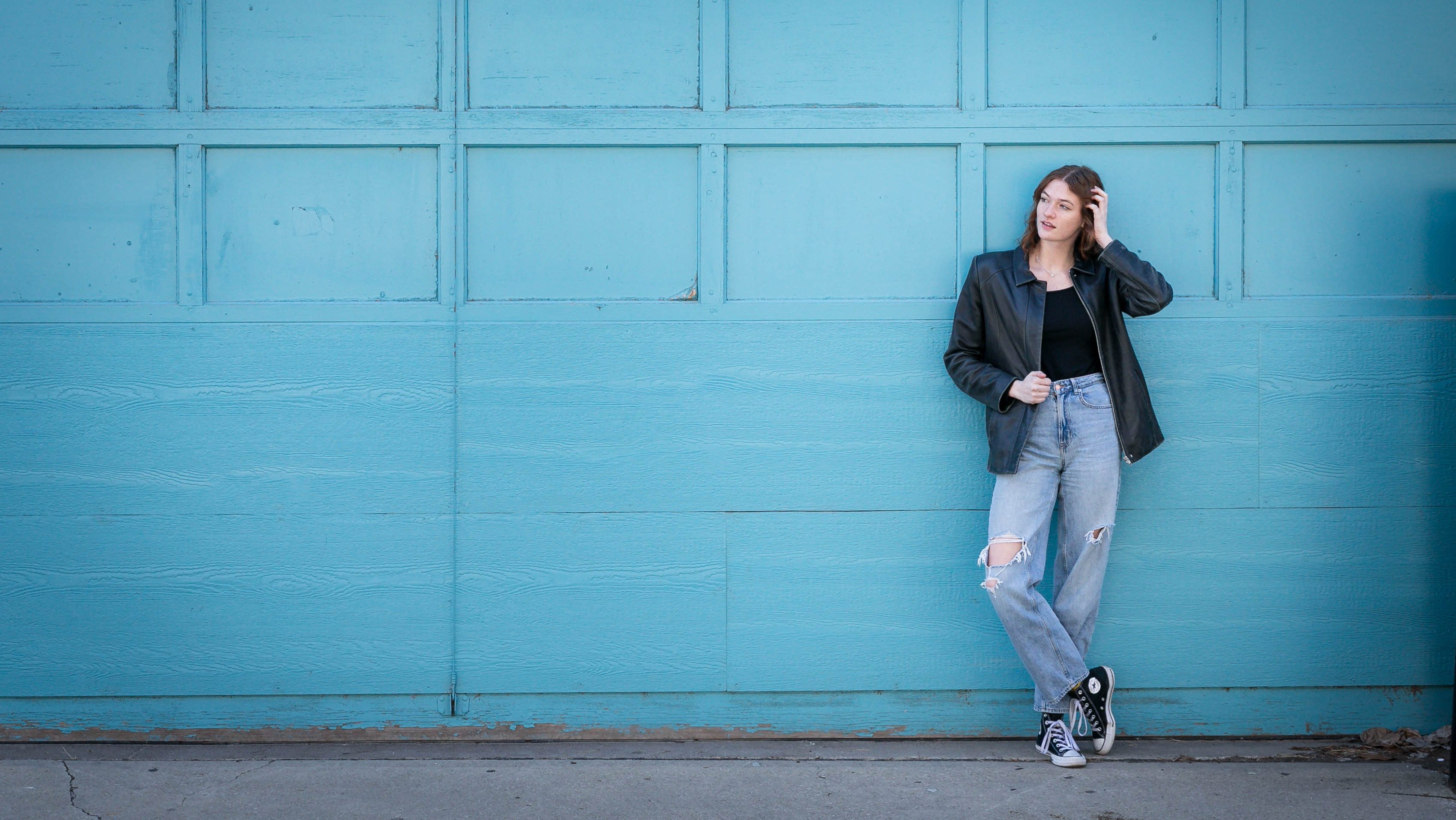 Woman in a black jacket and ripped jeans leans against a bright teal garage door, looking thoughtful. She stands on a concrete floor.