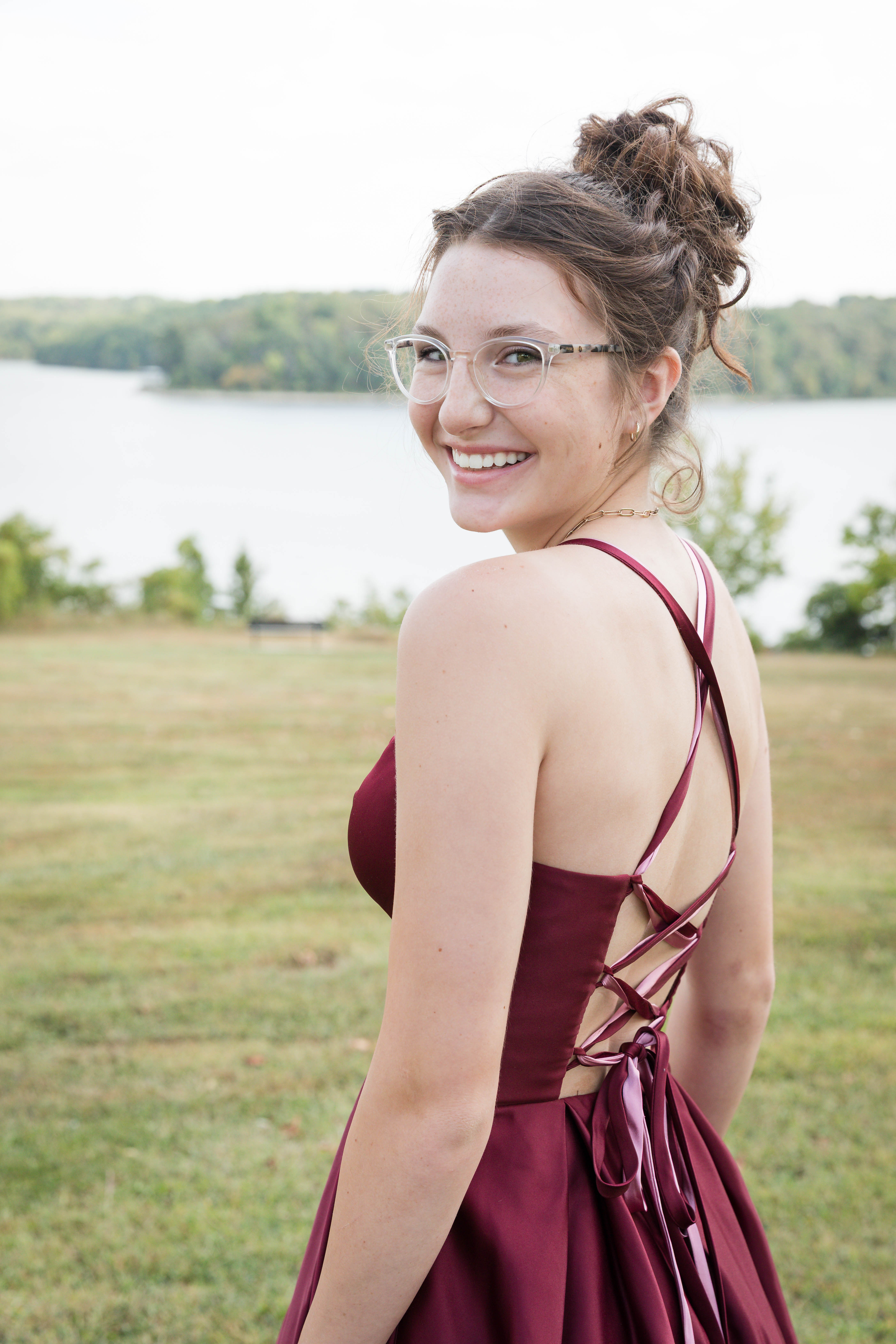 Smiling woman in a burgundy dress stands in a grassy field with a lake and trees in the background under a clear sky.