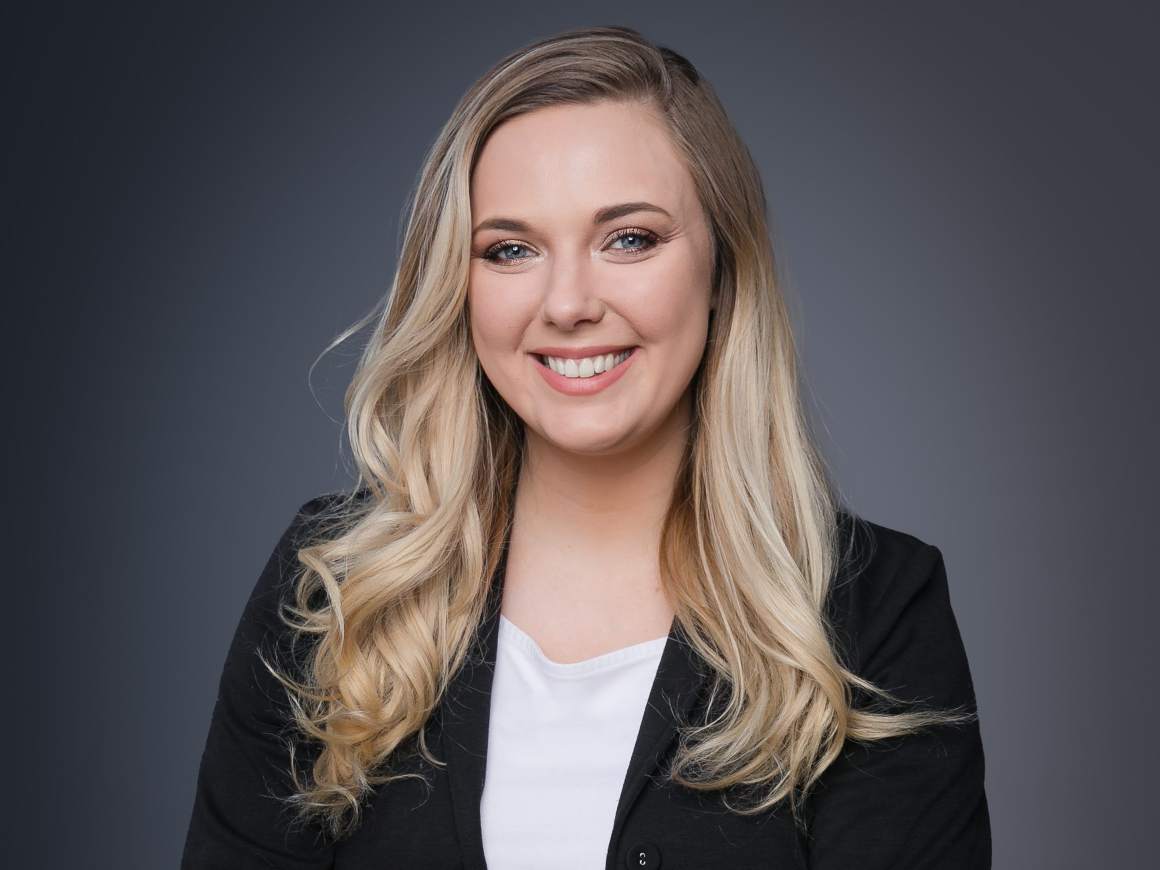 Smiling woman with long blonde hair in a black blazer on a gray gradient background. She exudes a confident and friendly demeanor.