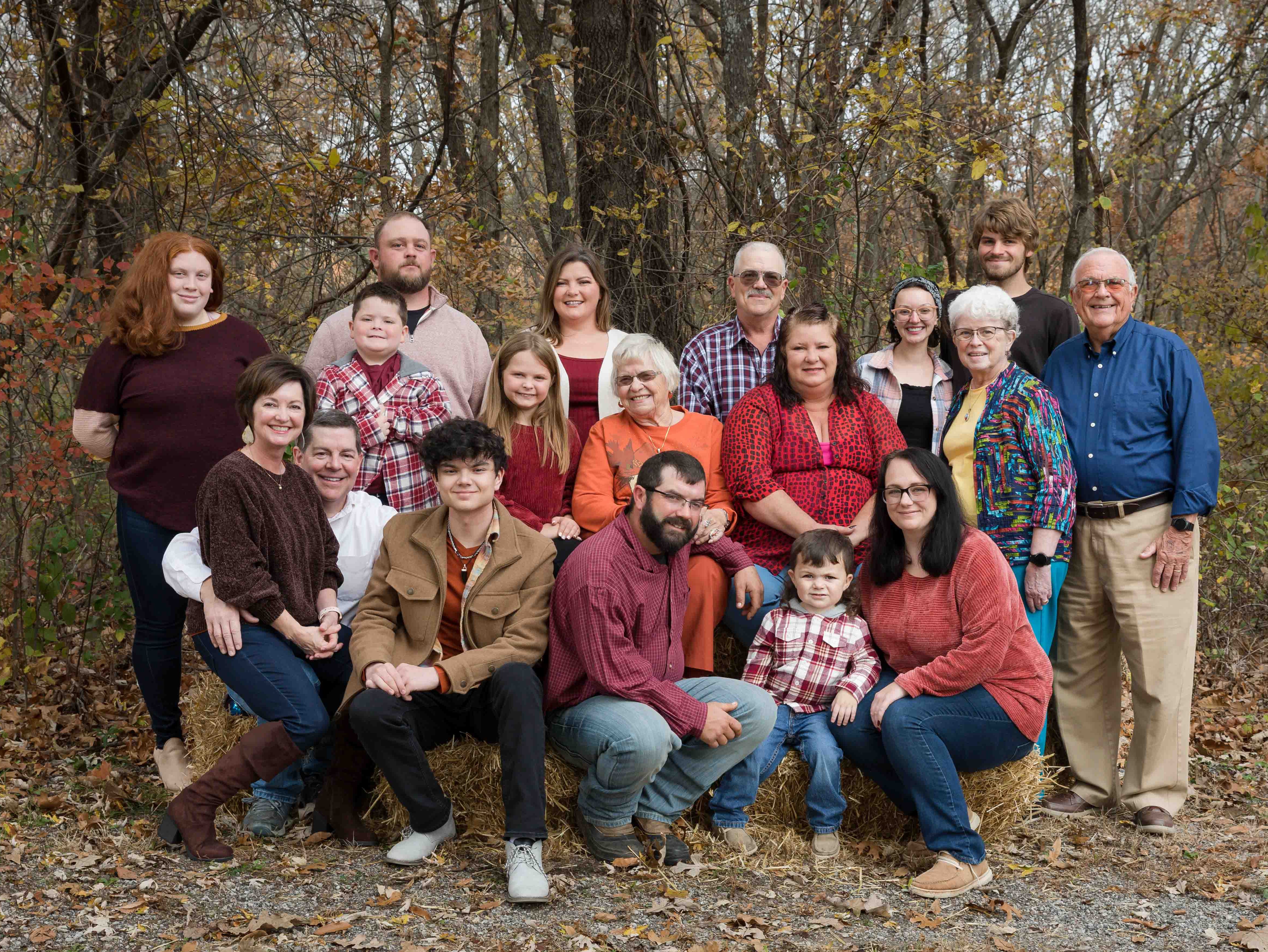 A large family poses outdoors on hay bales, surrounded by autumn trees. They wear casual, colorful clothing and smile warmly at the camera.