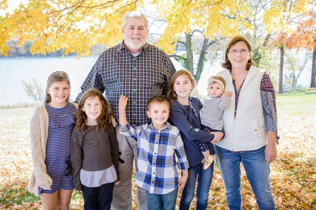 Family of seven smiling under autumn leaves by a lake. Adults stand at the ends, children between them. Bright colors, joyful mood.