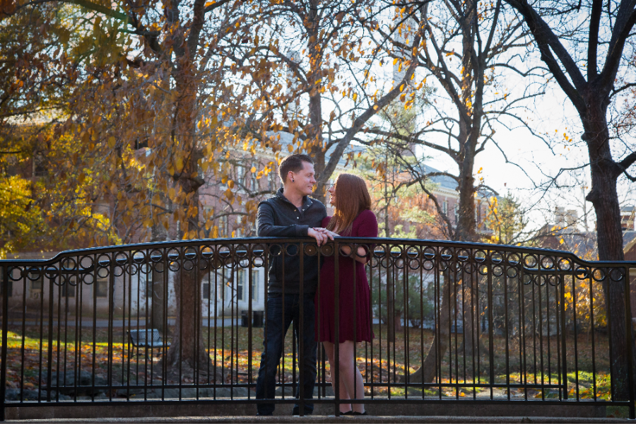 A couple stands on a black wrought iron bridge in a park with autumn trees. They are smiling and holding hands, with sunlight filtering through.