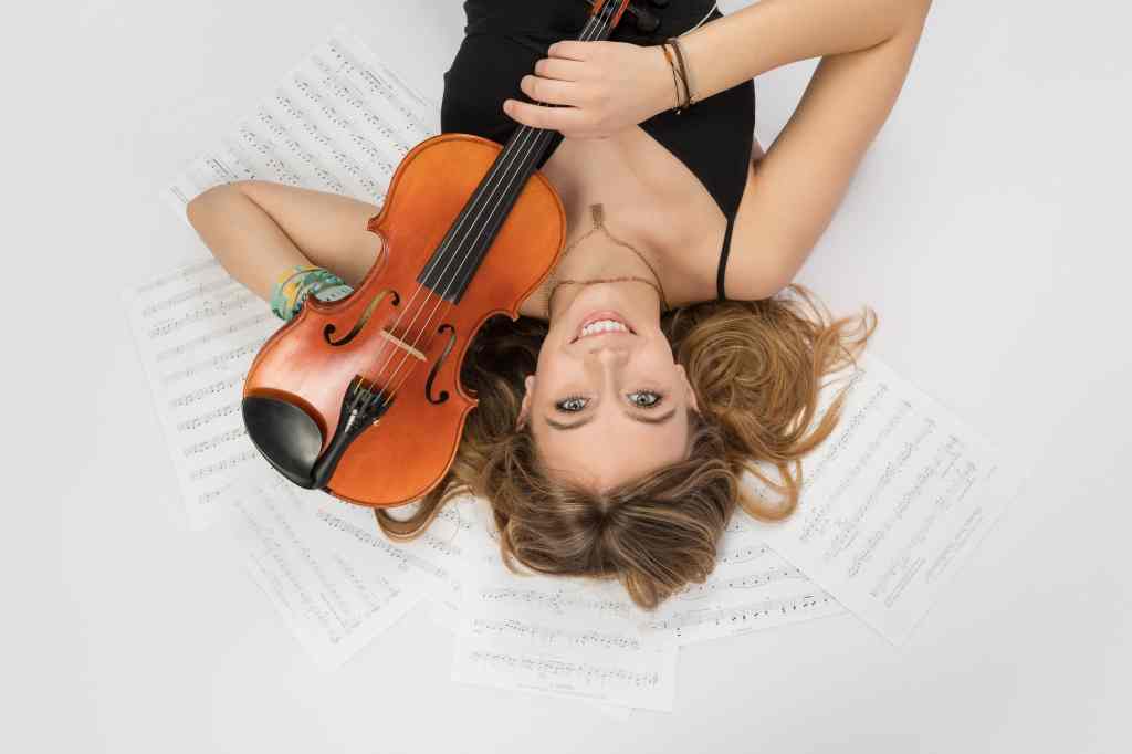 A woman lies on sheet music, smiling, holding a violin. She's wearing a black dress. The scene is bright and playful.