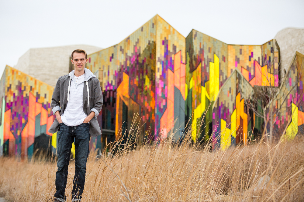 Person in casual clothes standing in front of a colorful, geometric-patterned building with tall grass in the foreground. Cloudy sky.