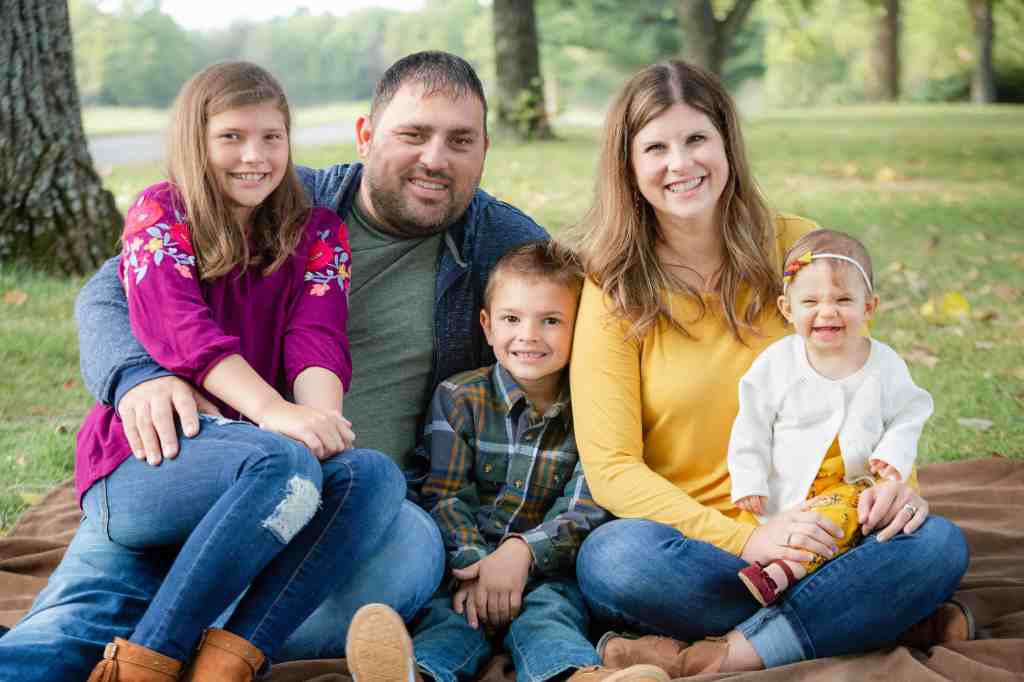 Smiling family of five sitting on a blanket in a park. Dad wears blue, mom in yellow, kids in bright outfits. Green trees in background.