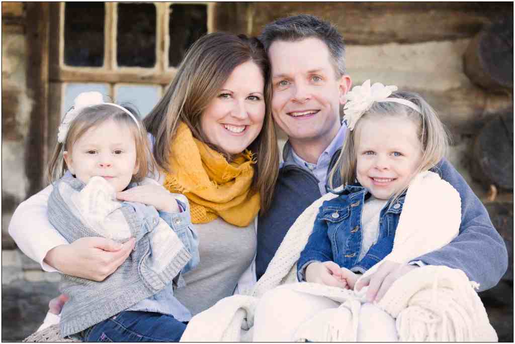 Smiling family of four sitting together in front of a rustic log cabin. The mother wears a yellow scarf, and the children have headbands.
