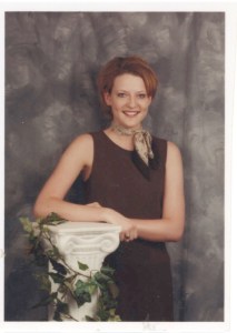 Smiling woman in brown dress with a scarf, leaning on a white column with ivy. Gray marbled background adds elegant contrast.