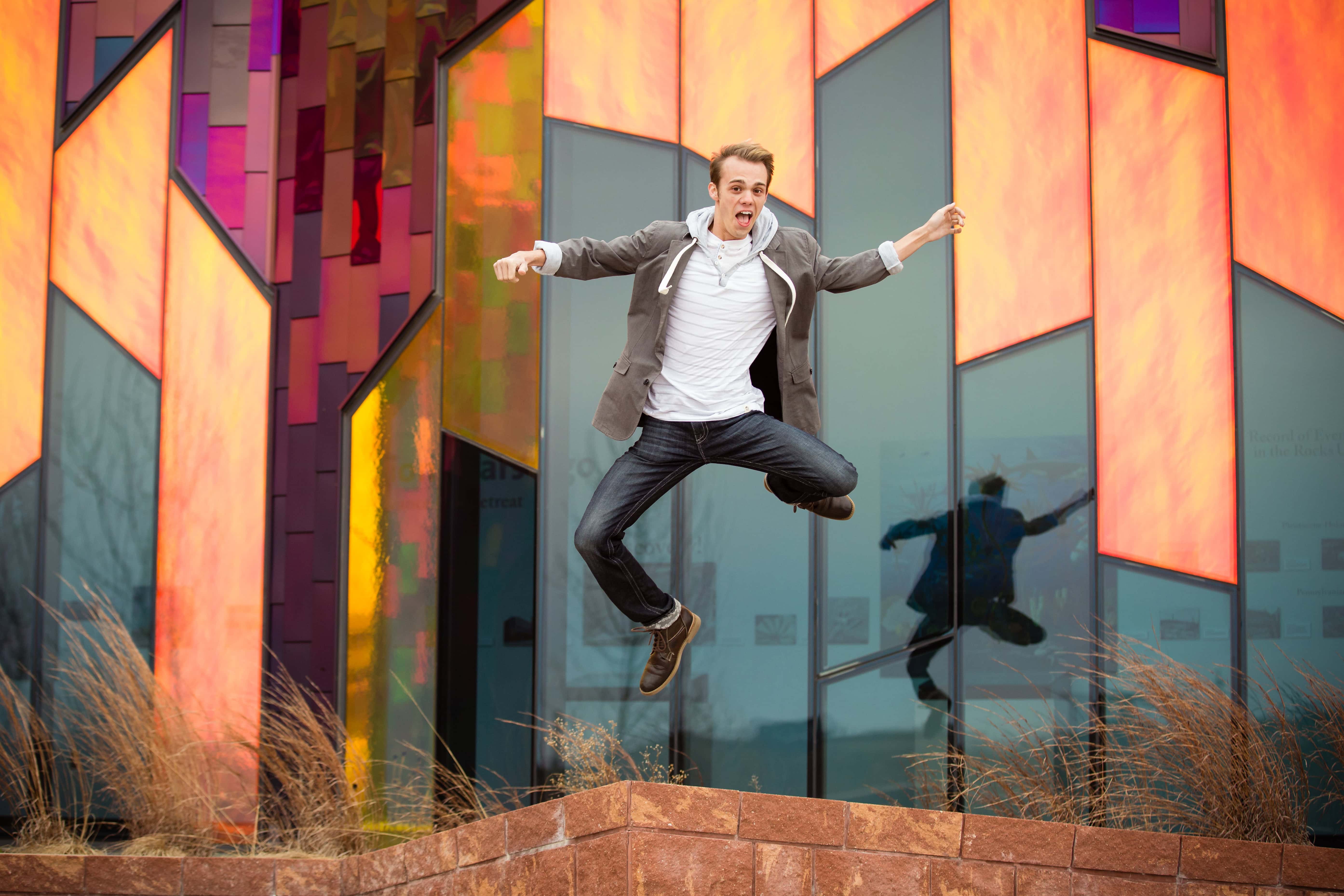A man jumps joyfully in front of a colorful glass building with orange and red panels, wearing jeans, a white shirt, and a gray jacket.