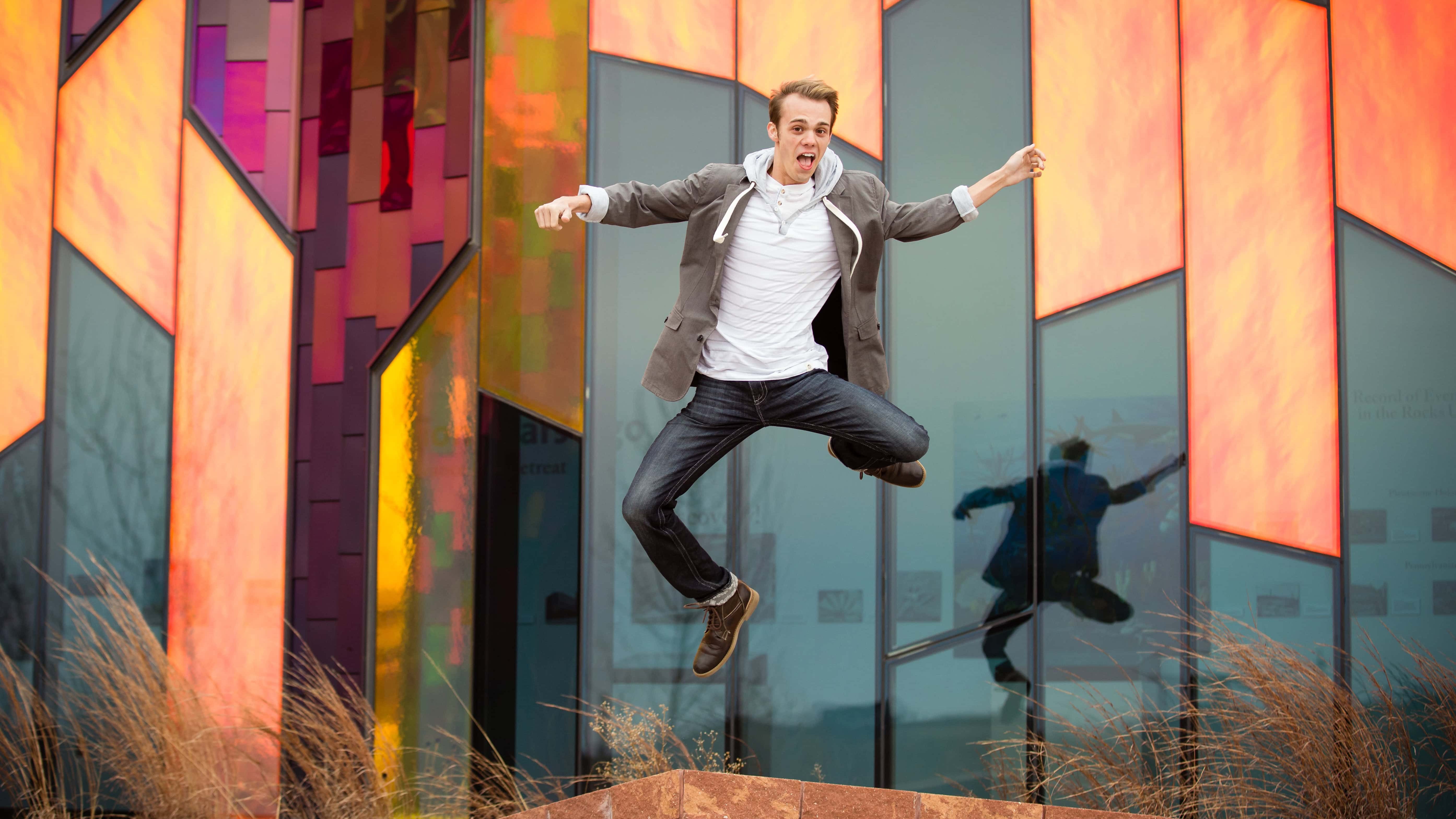 A man jumps joyfully in front of a colorful glass building with orange and red panels, wearing jeans, a white shirt, and a gray jacket.